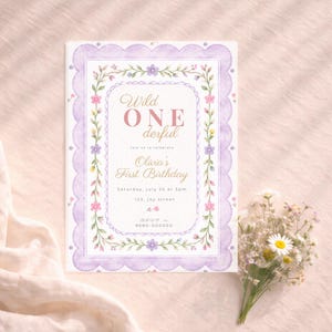 Pode incluir: Um convite de aniversário com tema floral com uma borda ondulada lavanda. O texto diz "Wild ONE derful" e "Olivia's First Birthday". O convite inclui a data, hora e local. Um pequeno buquê de flores brancas.