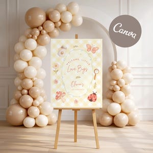 Op de afbeelding: Een houten ezel toont een welkomstbord met de tekst "Welcome, Little Love Bugs" en "Olivia's 1st Birthday". Het bord heeft een gele ruitjesachtergrond met illustraties van een vlinder, lieveheersbeestje en paddenstoel. Beige ballonnen vormen een boog en een kolom.