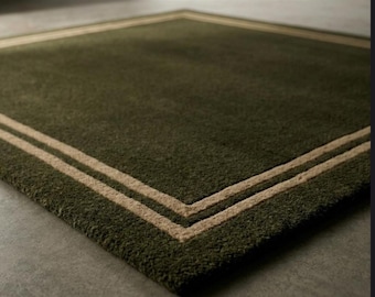May include: A square, olive green rug with a double border in a lighter tan color. The rug has a plush texture and is displayed on a gray surface. The border frames the rug's edges.