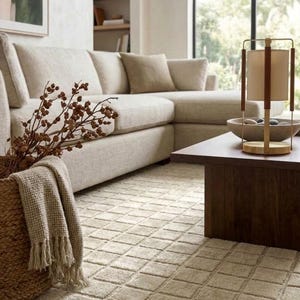 May include: A beige sectional sofa with matching throw pillows, a woven basket containing dried branches and a textured throw blanket, a dark wood coffee table, and a cylindrical lamp with a light-coloured shade and a wooden frame.