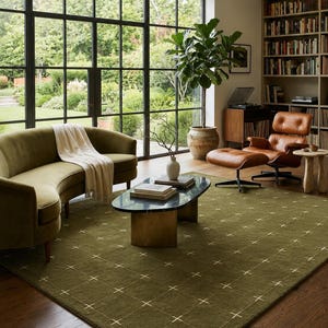 May include: A living room scene featuring a green rug with a cross pattern, a curved green sofa, and a brown leather chair. A coffee table with a black marble top and a large window overlooking a garden are also visible.