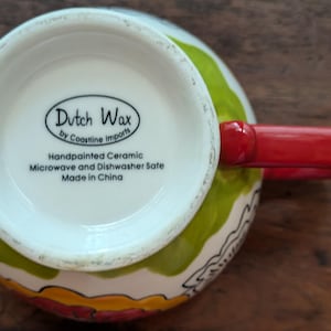 May include: A white ceramic mug with a green, yellow, and red floral design. The mug has a red handle and says "Dutch Wax by Coastline Imports" on the bottom. It is handpainted and microwave and dishwasher safe.