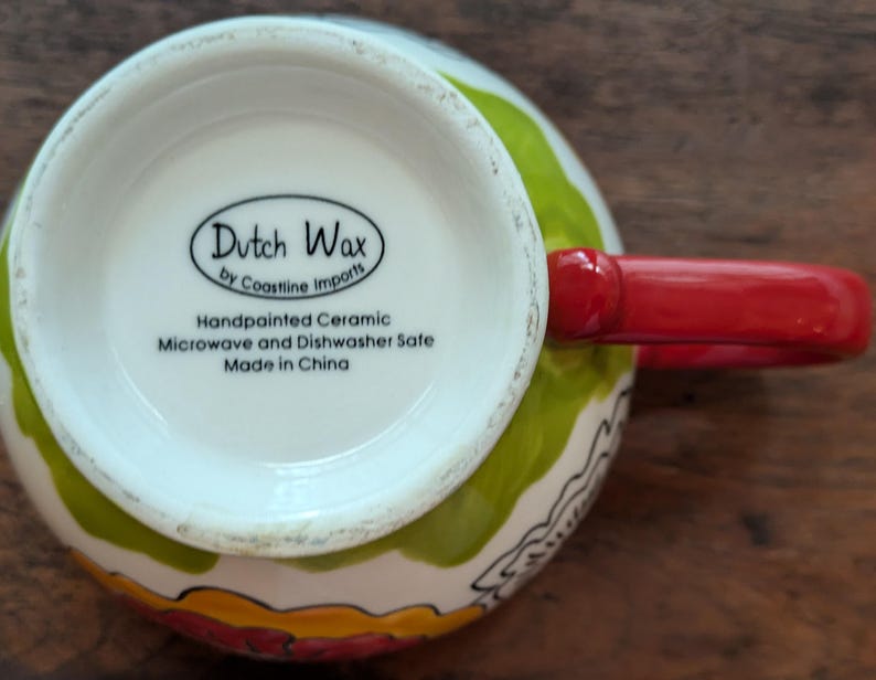 May include: A white ceramic mug with a green, yellow, and red floral design. The mug has a red handle and says "Dutch Wax by Coastline Imports" on the bottom. It is handpainted and microwave and dishwasher safe.