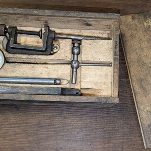 May include: A wooden box with a hinged lid, containing a dial gauge, a clamp, and a metal rod. The box is open and the contents are visible.