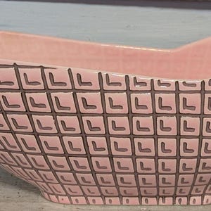 May include: A pink ceramic decorative bowl with a geometric pattern. The bowl has a boat-like shape with a wide opening and a textured surface. The exterior features a repeating square design in a darker shade of pink, creating a contrast against the lighter pink background.