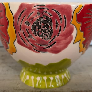 May include: A white ceramic bowl with a green base and a floral design. The bowl features red, yellow, and black flowers with a white background.