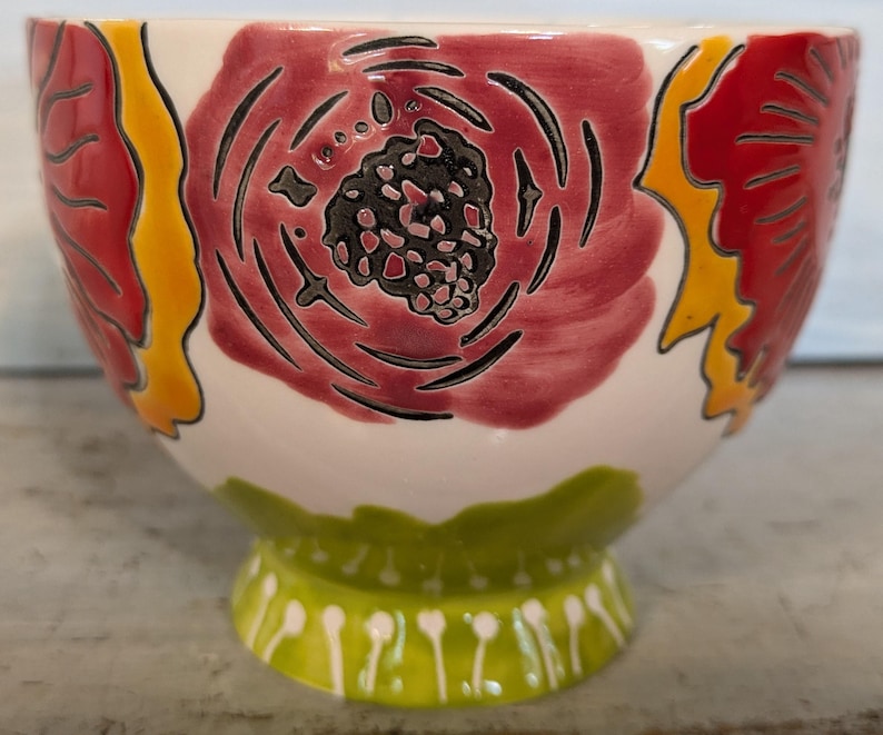 May include: A white ceramic bowl with a green base and a floral design. The bowl features red, yellow, and black flowers with a white background.