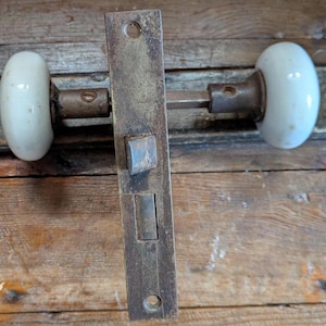 May include: Antique door hardware set featuring a rectangular, aged metal plate with a latch and two white oval porcelain doorknobs. The hardware shows signs of wear and age, with a rustic, vintage aesthetic.