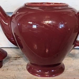 May include: A burgundy ceramic teapot with a matching lid. The teapot has a curved spout, a large handle, and a rounded body. The lid sits to the left of the teapot. The teapot is a classic design, suitable for serving tea.