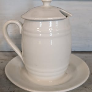May include: A white ceramic sugar bowl with a lid and a small handle, resting on a matching saucer. The sugar bowl has a curved handle and a small opening in the lid for a spoon. The design is simple and classic.