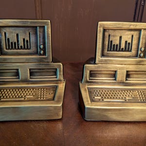 May include: A pair of gold-toned computer-shaped bookends. Each bookend features a monitor, a keyboard, and a hard drive. The bookends are a decorative way to hold books or other items on a shelf.
