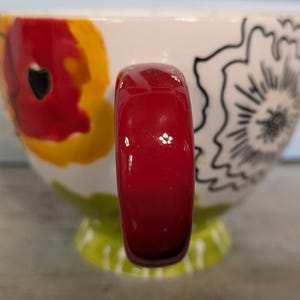May include: A white ceramic mug with a red handle. The mug has a floral design with red, yellow, and black accents.