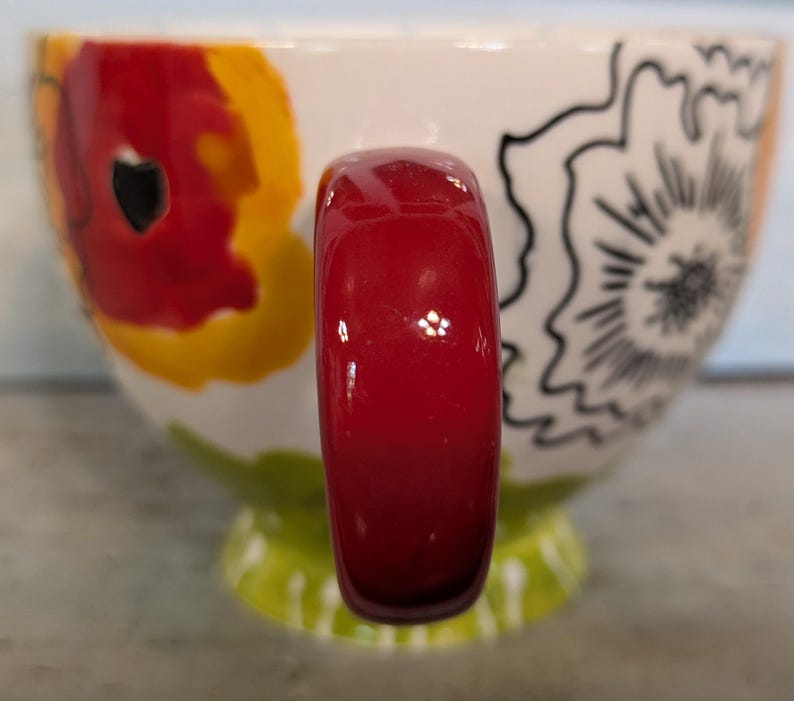 May include: A white ceramic mug with a red handle. The mug has a floral design with red, yellow, and black accents.
