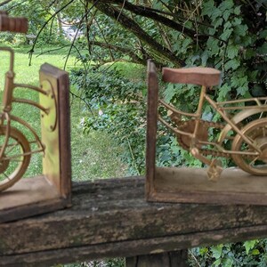 May include: A pair of bookends featuring a gold-toned metal bicycle design split into two sections. Each piece is mounted on a rectangular wooden base, with the front section showing the handlebars and wheel, and the rear section showing the seat and wheel.