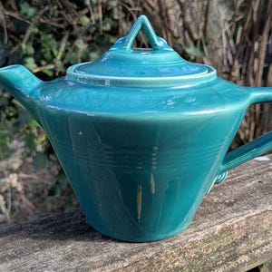 May include: A teal ceramic teapot with a matching lid. The teapot has a handle and a spout. The lid has a small loop for easy removal. The teapot is sitting on a wooden surface.