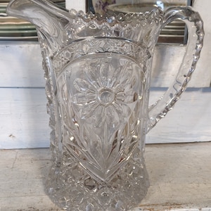 May include: A clear glass pitcher with a floral design. The pitcher has a wide mouth and a handle. The design is intricate and detailed, with a large flower in the center.