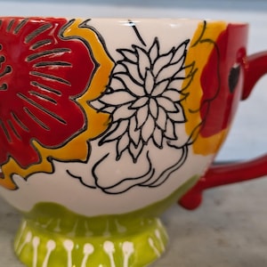May include: A white ceramic mug with a red, yellow, and black floral design. The mug has a red handle and a green base with white dots.
