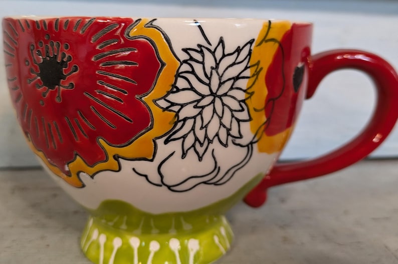 May include: A white ceramic mug with a red, yellow, and black floral design. The mug has a red handle and a green base with white dots.