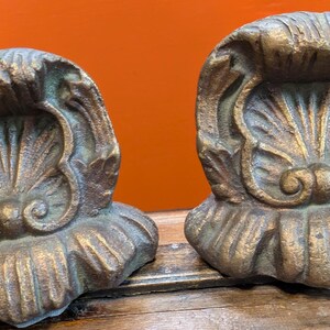 May include: A pair of heavy, metallic bookends featuring a raised, ornate shell motif with a weathered gold and bronze finish. Each piece displays intricate, symmetrical scrollwork and fluted detailing, showcasing a textured, antique-style surface.