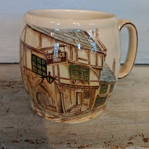 May include: A ceramic mug with a brown and white illustration of a building with the text "George Inn" on a sign. The mug has a handle and is a light brown color.