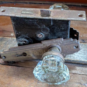 May include: An antique door lock set with a clear glass doorknob and a dark metal lock mechanism. The set includes a rectangular faceplate and a matching strike plate, all showing signs of age and wear, resting on a wooden surface.