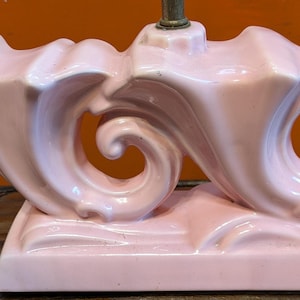May include: A pink ceramic lamp base with a unique, swirling design. The base features two large, curved elements with a scroll-like shape, connected by a central support. The lamp base sits on a rectangular platform and has a brass fitting for the lamp.