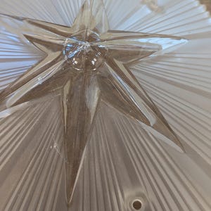 May include: A clear glass light fixture shade featuring a prominent central star design with raised, radiating rays extending to the scalloped edges. The glass has a frosted finish and includes two circular mounting holes for installation.