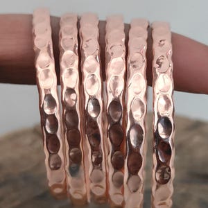May include: Six metallic copper bangles arranged in a row, each featuring a distinct hammered texture with reflective, circular indentations along the surface. The text silversanthoAtoZ is printed in yellow at the bottom of the image.