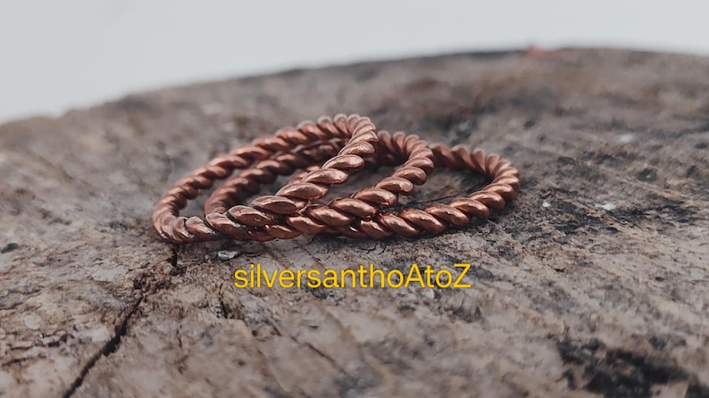 Op de afbeelding: Drie koperkleurige, gedraaide ringen in touwstijl worden getoond op een getextureerd, bruin houten oppervlak. De ringen zijn scherp, wat hun ingewikkelde gevlochten ontwerp laat zien. De ringen hebben een warme, roodbruine kleur.