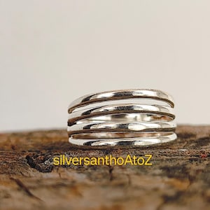 May include: A close-up of a silver ring with a multi-layered design. The ring has a textured surface and is made of a shiny, metallic material. The text "silversanthoAtoZ" is visible on the wooden surface.