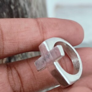 Puede incluir: Un anillo de plata con un dise&ntilde;o de cruz. El anillo tiene una banda gruesa y una forma de cruz tridimensional en el centro. El metal tiene una superficie pulida y reflectante, y el anillo parece hecho a mano.