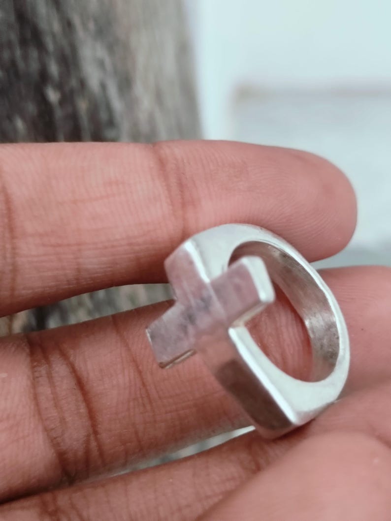 Puede incluir: Un anillo de plata con un dise&ntilde;o de cruz. El anillo tiene una banda gruesa y una forma de cruz tridimensional en el centro. El metal tiene una superficie pulida y reflectante, y el anillo parece hecho a mano.
