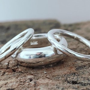 Puede incluir: Tres anillos de plata se muestran sobre una superficie marrón texturizada. Dos anillos más finos se colocan alrededor de un anillo más grueso y redondeado. Los anillos son reflectantes, con un acabado pulido, y parecen ser de metal.