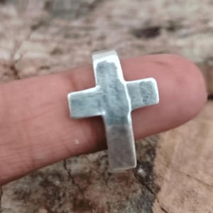 以下が含まれることがあります： シンプルでクラシックなデザインのシルバーカラーのクロス ペンダント。十字架は指で支えられており、そのサイズと形状を示しています。ペンダントはわずかにテクスチャ加工された表面を持っています。