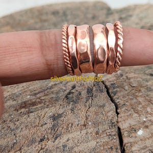 May include: A set of copper rings displayed on a finger. The rings have various designs, including hammered and twisted rope textures. The rings are a warm copper color, and the image is taken in natural light.
