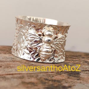 Pode incluir: Um anel largo prateado com um design detalhado de abelha. O anel apresenta uma banda texturizada com padrões florais e uma abelha proeminente no centro. O texto "silversanthoAtoZ" está impresso em amarelo na parte inferior.
