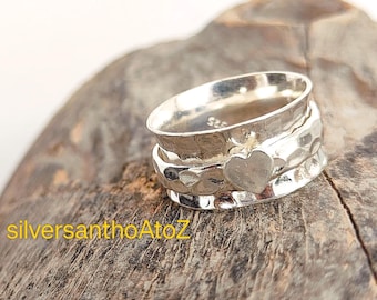 Può includere: Anello spinner argentato con un ciondolo a forma di cuore. L'anello presenta una texture martellata e il testo "silversanthoAtoZ" in giallo. È appoggiato su una superficie di legno.