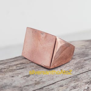 Pode incluir: Um anel cor de cobre com uma face retangular e um design geométrico. O anel está sobre uma superfície de madeira. O texto "silversanthoAtoZ" é visível.