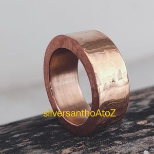 Puede incluir: Un anillo ancho, de color cobre, con una superficie lisa y pulida. El anillo tiene un diseño sencillo de banda. El texto "silversanthoAtoZ" es visible en el anillo. Este anillo es una joya.