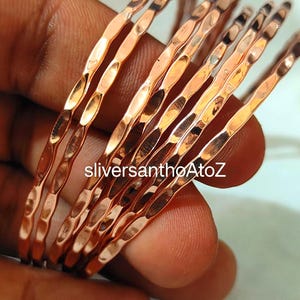 May include: A collection of copper-coloured bangles with a hammered texture. The bracelets are held in a hand, and the surface reflects light. The text "sliversanthoAtoZ" is visible on the image.