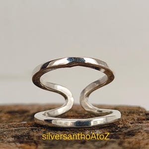 Puede incluir: Un anillo de plata de doble banda con un diseño abierto único. El anillo presenta dos bandas curvas conectadas por una banda superior, creando un aspecto moderno y elegante. El anillo se muestra sobre una superficie de madera. El texto "silversanthoAtoZ" es visible.