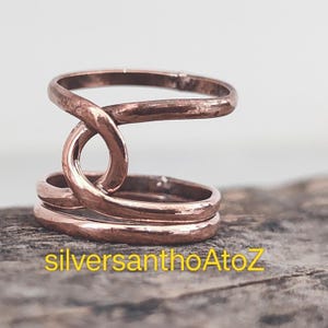 May include: A copper-colored ring with a unique design. The ring features a looped central element that intersects with the band. The band is composed of multiple parallel rings. The text "silversanthoAtoZ" is visible at the bottom.