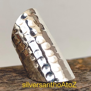 Peut inclure: Une bague en argent avec une texture martelée. La bague a un design incurvé et allongé et une surface brillante et réfléchissante. La bague est présentée sur une surface en bois. Le texte "silversanthoAtoZ" est visible en bas.
