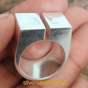 Puede incluir: Un anillo de plata con un diseño moderno. El anillo presenta una banda gruesa con dos bloques rectangulares separados por un espacio. El anillo está hecho de un material metálico brillante. El texto "silversanthoAtoZ" es visible en el anillo.