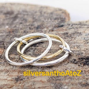 Puede incluir: Tres anillos entrelazados, dos plateados y uno dorado, descansan sobre una superficie marrón texturizada. Los anillos tienen un diseño liso y sencillo, con una ligera curva. El texto "silversanthoAtoZ" es visible en amarillo.