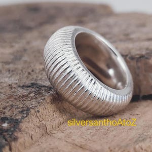 Könnte beinhalten: Ein silberner Ring mit einer strukturierten, geriffelten Oberfläche. Der Ring ist dick und abgerundet, mit poliertem Innenraum. Der Ring wird auf einer rauen Holzoberfläche präsentiert. Der Text "silversanthoAtoZ" ist sichtbar.