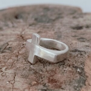 Puede incluir: Un anillo plateado con un dise&ntilde;o de cruz. El anillo presenta una banda gruesa y una cruz en relieve. El anillo est&aacute; hecho de un material met&aacute;lico brillante y se encuentra sobre una superficie marr&oacute;n texturizada.