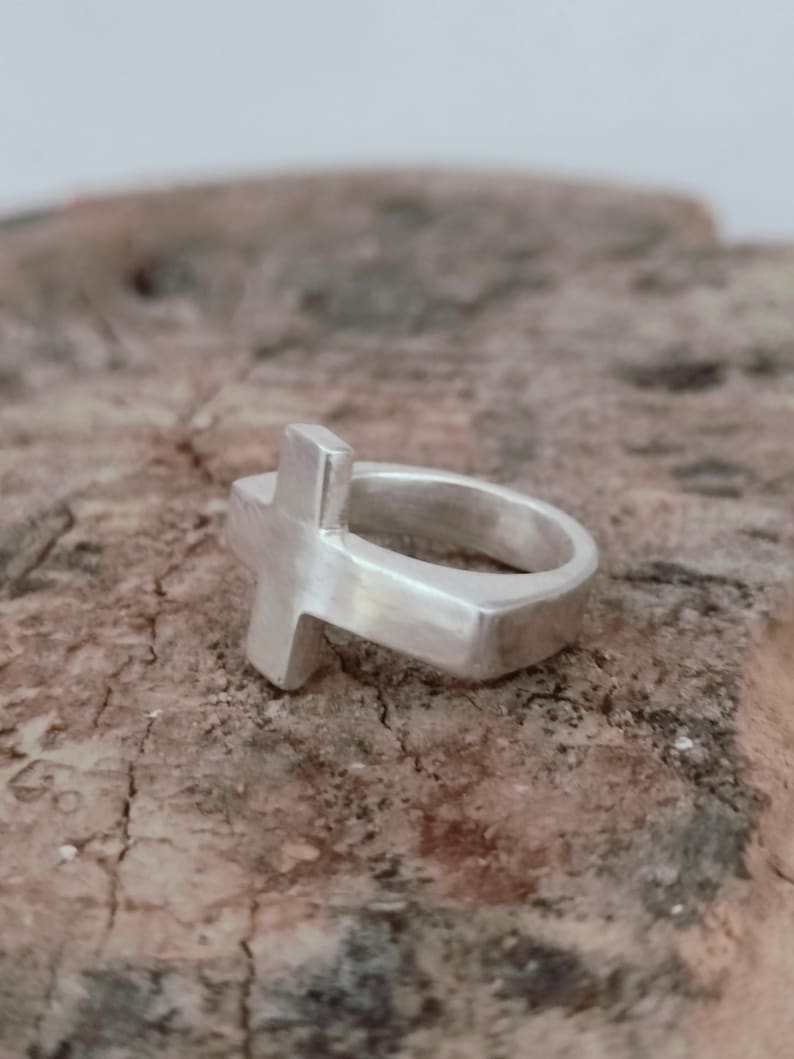 Puede incluir: Un anillo plateado con un dise&ntilde;o de cruz. El anillo presenta una banda gruesa y una cruz en relieve. El anillo est&aacute; hecho de un material met&aacute;lico brillante y se encuentra sobre una superficie marr&oacute;n texturizada.