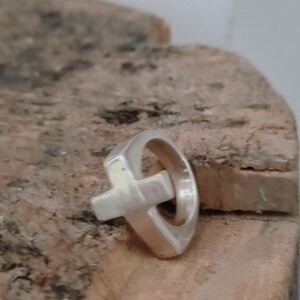 Puede incluir: Un anillo de plata con un dise&ntilde;o de cruz. El anillo presenta una banda redondeada con una forma de cruz adjunta. El anillo est&aacute; sobre una superficie texturizada de color marr&oacute;n claro.