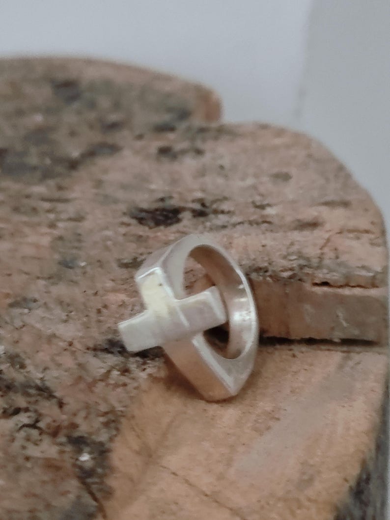 Puede incluir: Un anillo de plata con un dise&ntilde;o de cruz. El anillo presenta una banda redondeada con una forma de cruz adjunta. El anillo est&aacute; sobre una superficie texturizada de color marr&oacute;n claro.